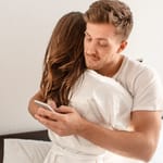 Nainen ja mies halaa, mies selaa puhelinta naisen selän takana samalla.